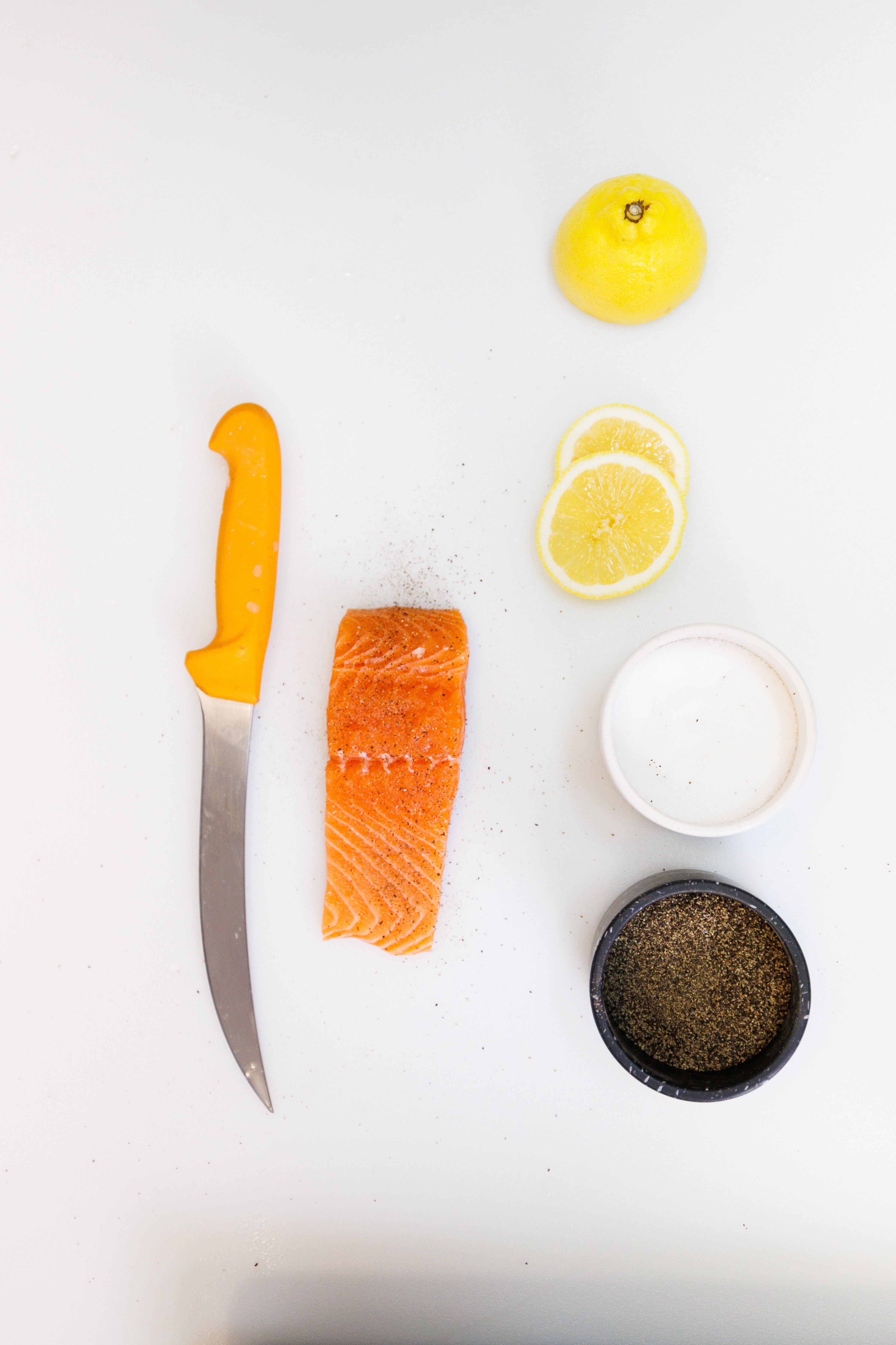 Six objects laid out on a white background - a knife, a piece of atlantic salmon, a lemon, lemon slices, salt, pepper.