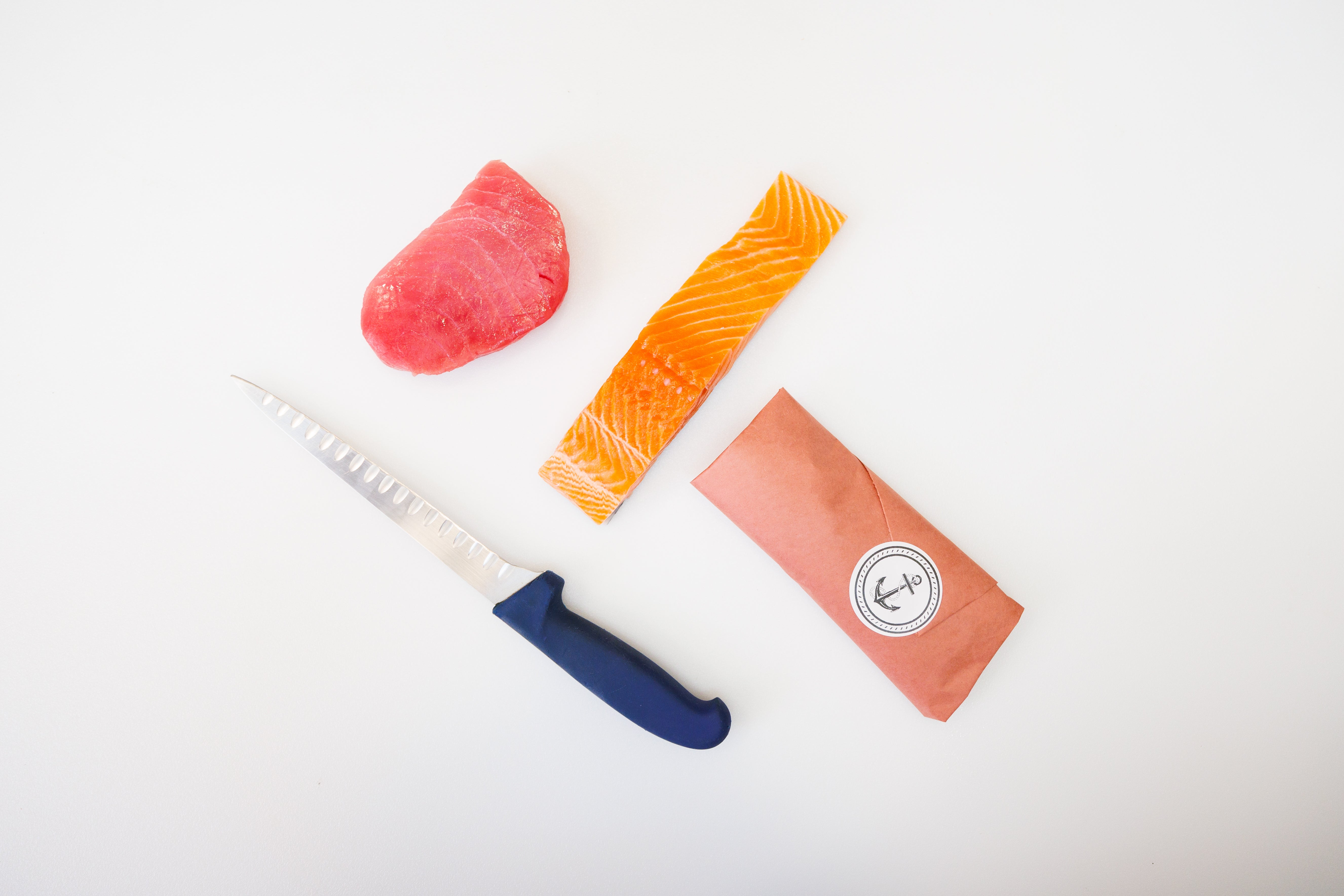 Four objects laid out on a white background - a knife, a piece of tuna, a piece of king salmon, a piece of fish wrapped in paper.