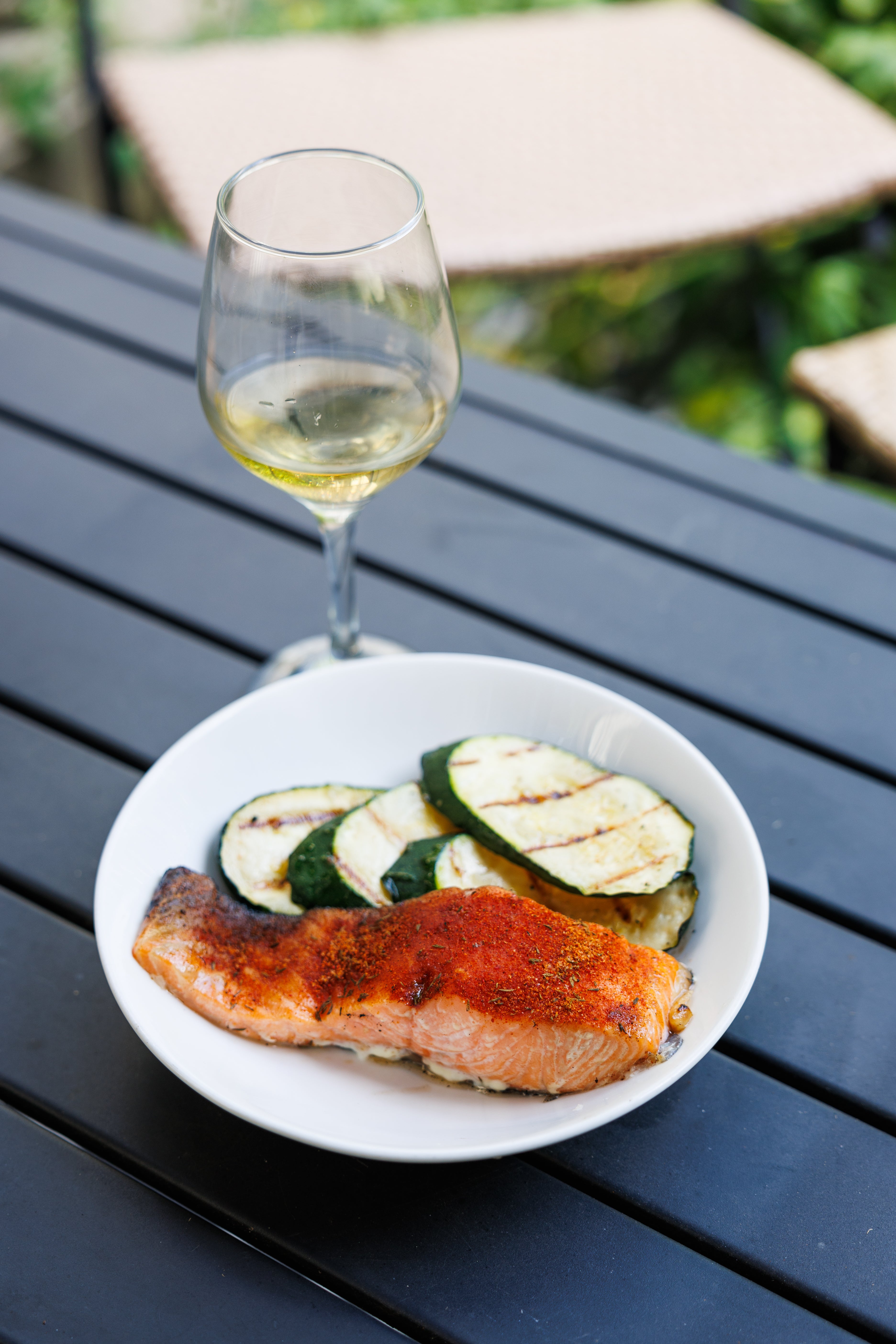 A cooked piece of salmon covered in red blackening rub, next to grilled zucchini slices and a glass of white wine