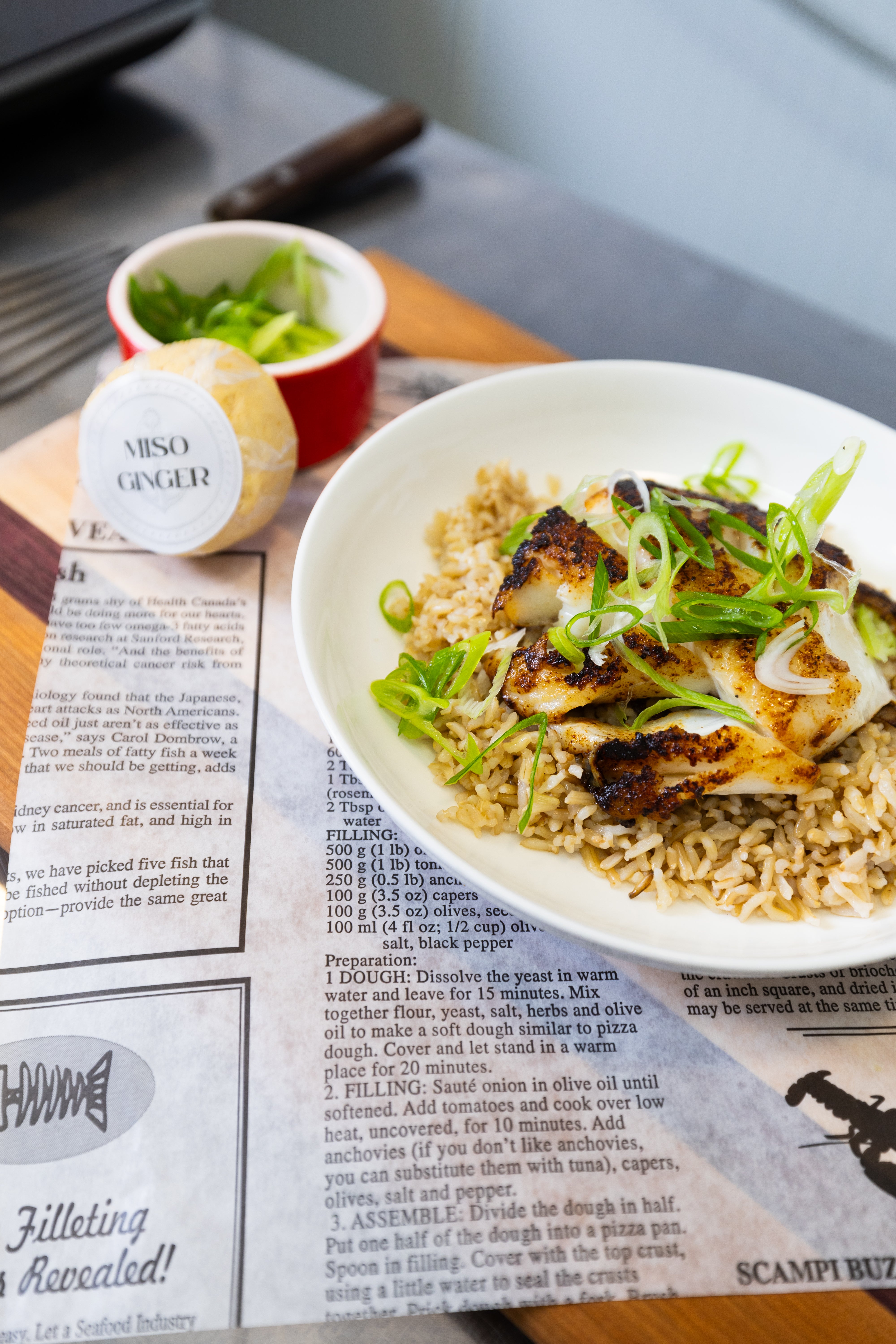 A plate of cooked fish on a bed of rice, next to a puck of miso ginger butter.