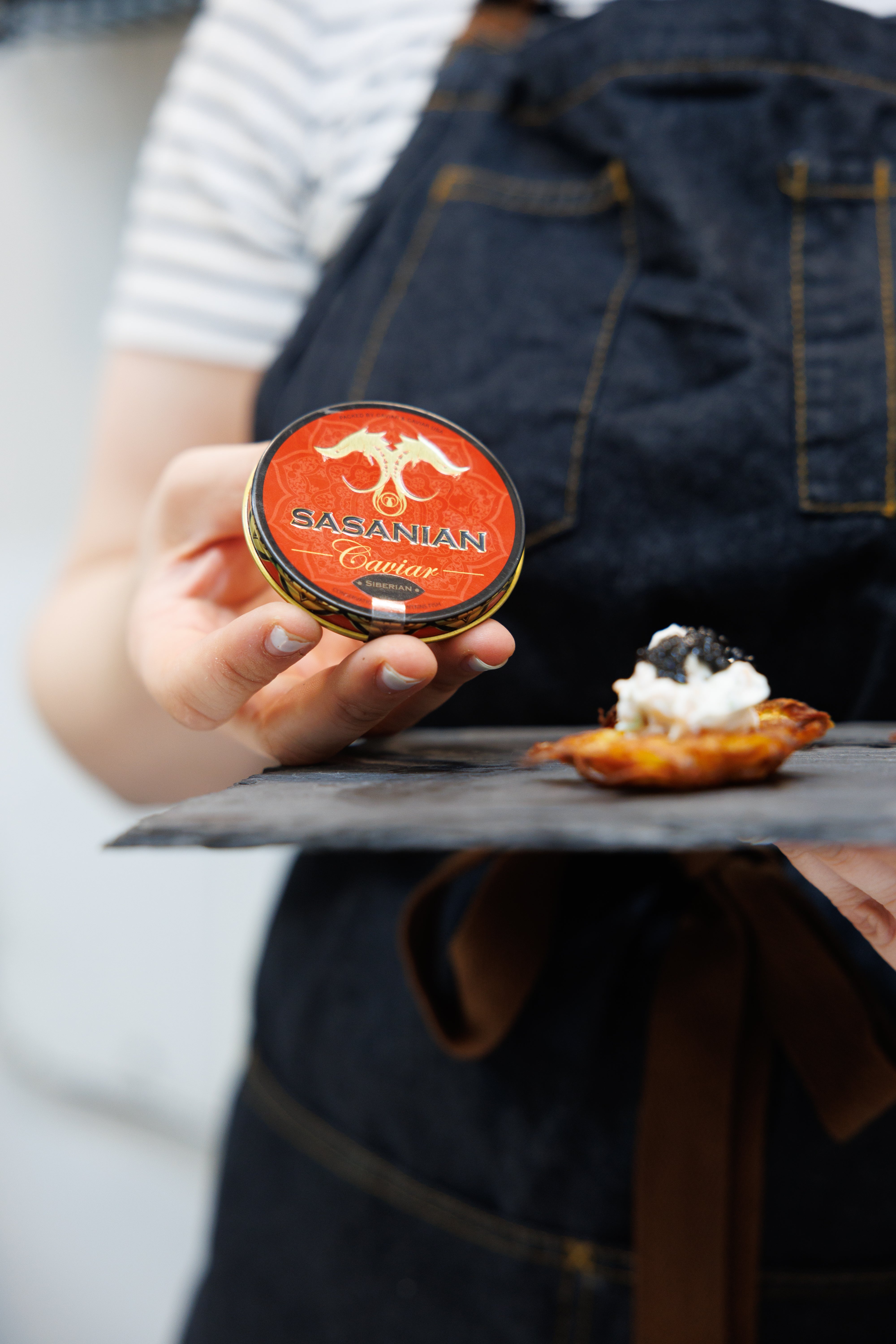 A hand holding a tin of Sasanian Siberian Caviar, with a suggested serving presentation