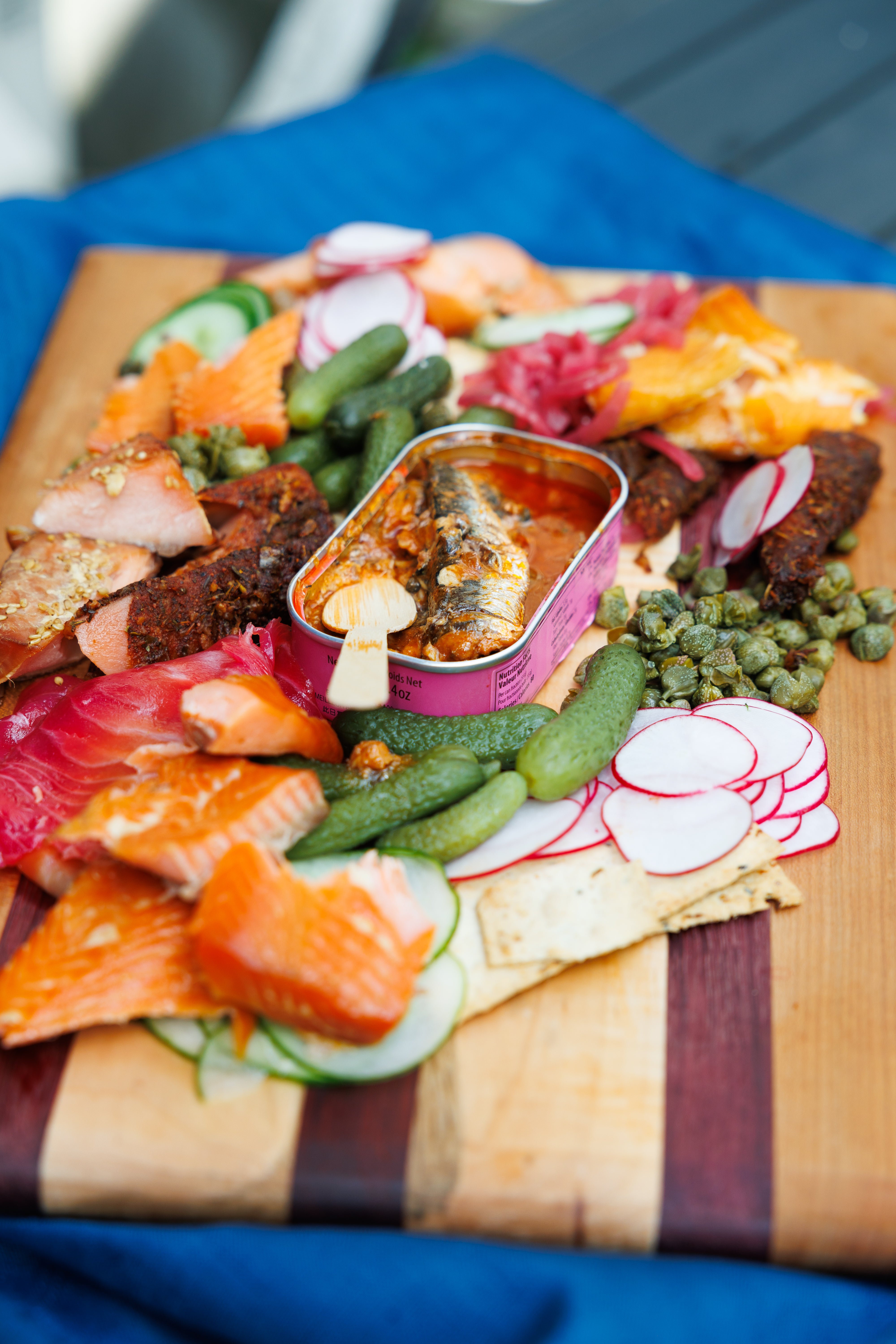 A tin of Porthos Sardines in Tomato Sauce, on a wooden board surrounded by smoked fish and garnish