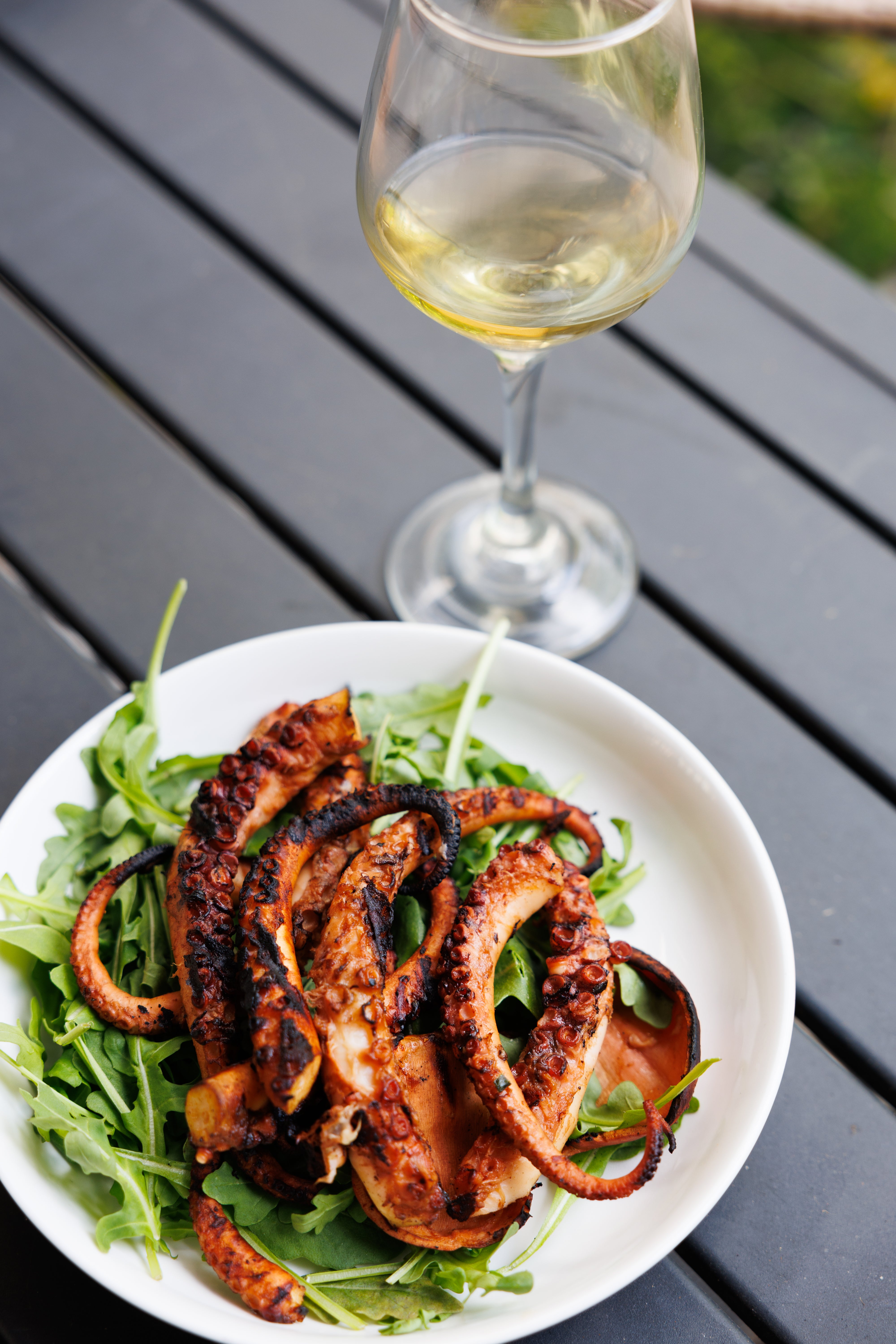 Grilled octopus on a bed of greens, next to a glass of white wine.