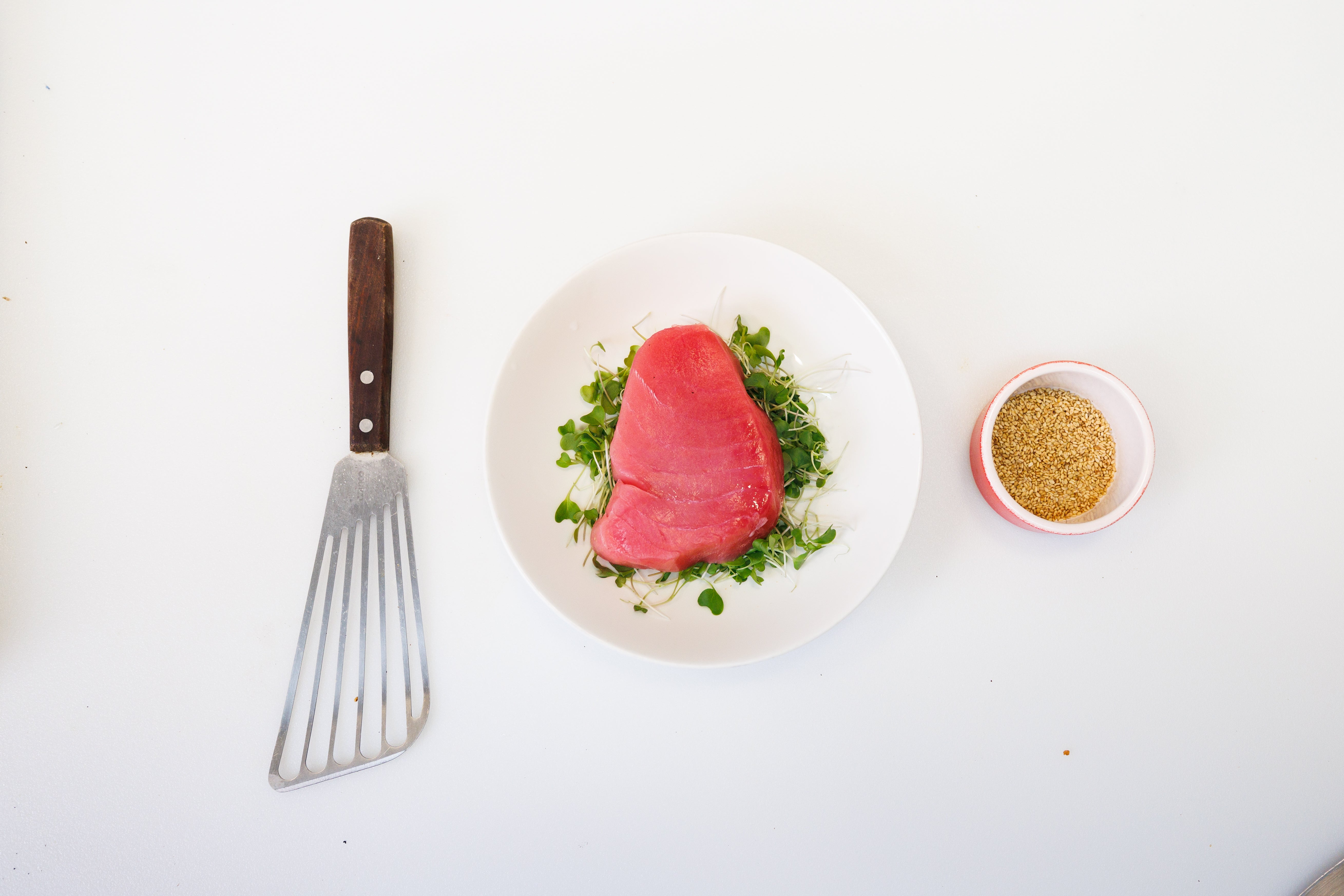 A tuna steak on a bed of greens next to a bowl of breadcrumbs and a flipping spatula